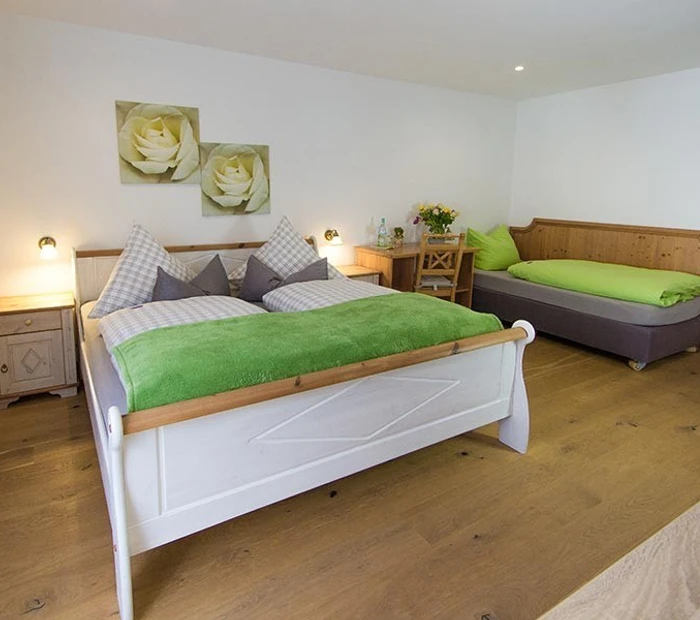 In dem Zimmer für 3 Personen stehen ein Doppelbett und eine Schlafcouch. Die Farben sind grau, grün, weiß. Der Fußboden ist aus Holzdielen.