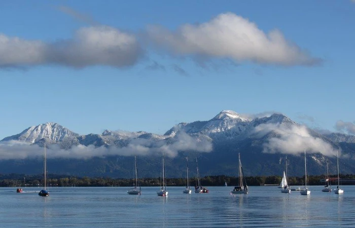 Blick vom Chiemsee auf Segelboote in die Chiemgauer Alpen. Die Berge sind mit Wolken umhüllt.