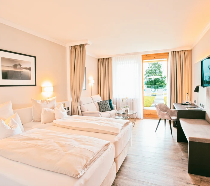 Sie blicken in ein Deluxe-Zimmer mit Doppelbett, Sofa und Blick auf den See vom Yachthotel Chiemsee.