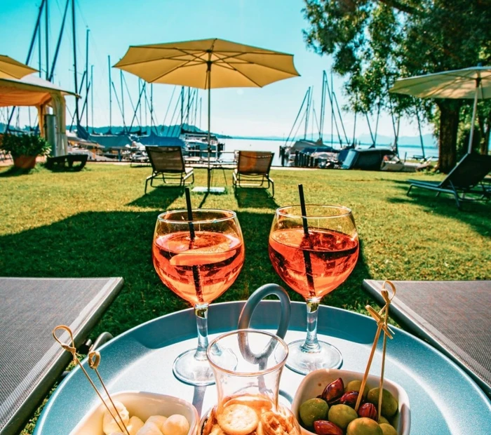 Im Gartenbereich mit Blick auf den Chiemsee stehen Aperol Spritz mit Knabereien auf einen kleinen Tisch.